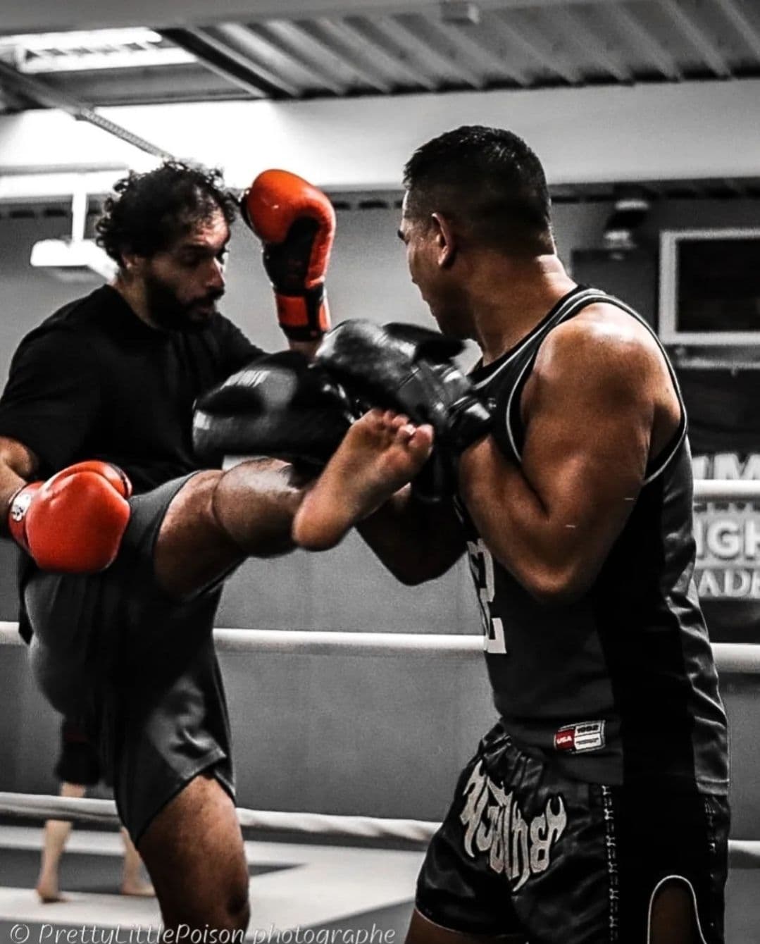 Deux pratiquants de Muay Thaï s’entraînent sur le ring, l’un réalisant un high kick tandis que l’autre bloque l’impact avec ses gants.